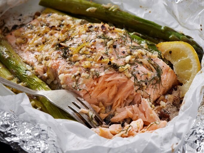 Ofenlachs mit grünem Spargel, Zitrone, Knoblauch und Dill in Backpapier und Alufolie mit einer Gabel. Draufsicht. | © Getty Images / LauriPatterson