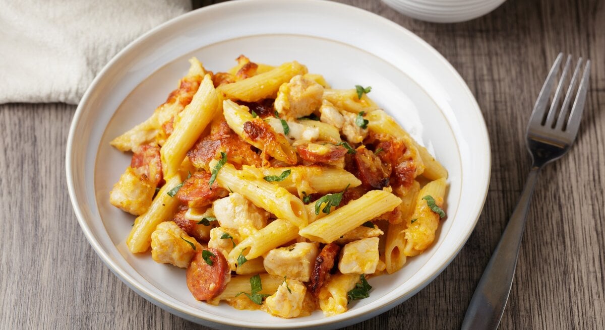 Pasta mit Bärlauch, Chorizo und Hähnchenbrust in weißem Pastateller auf Holz. Daneben eine Schale mit Bärlaich, ein Tuch und eine Gabel. Draufsicht.