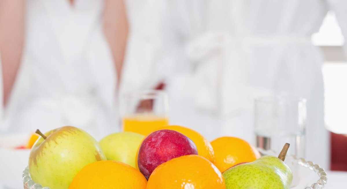 Obstschale mit Äpfeln, Birne, Pflaume und Mandarinen vor zwei Erwachsenen in weißen Bademänteln am Frühstückstisch.