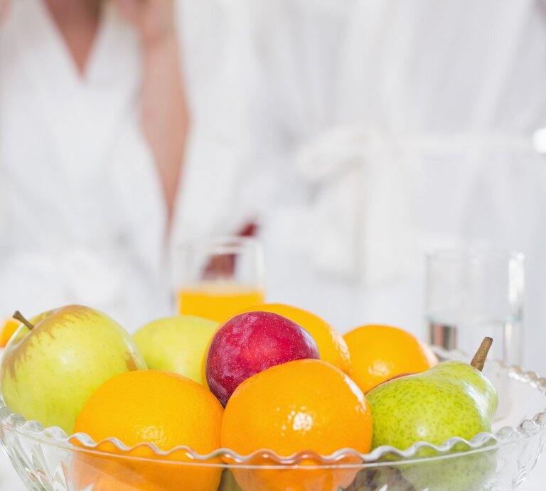Obstschale mit Äpfeln, Birne, Pflaume und Mandarinen vor zwei Erwachsenen in weißen Bademänteln am Frühstückstisch. | © Adobe Stock / WavebreakMediaMicro