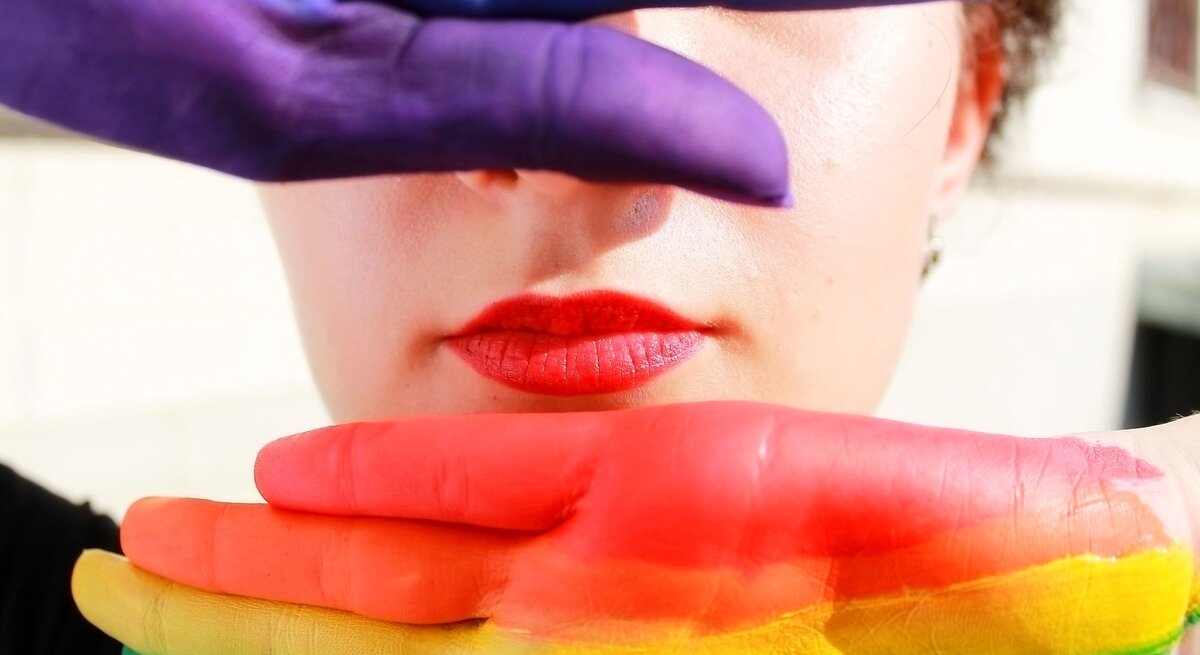 Junge Frau mit roten Lippen, deren Gesicht teils von zwei bunt bemalten Händen in Regenbogenfarben verdeckt wird.