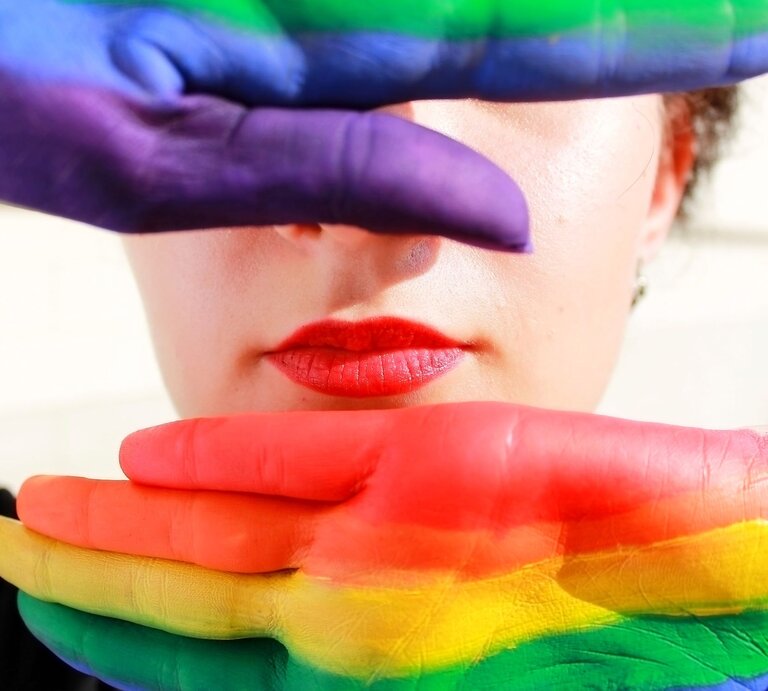 Junge Frau mit roten Lippen, deren Gesicht teils von zwei bunt bemalten Händen in Regenbogenfarben verdeckt wird. | © Adobe Stock / stasenso