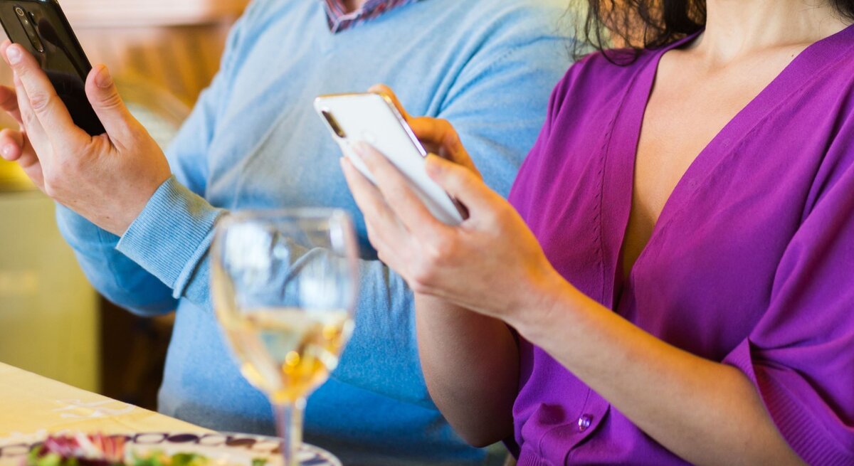 Eine Frau in lila Kleid und ein Mann in blauem Pulli starren beim Essen nebeneinander auf ihre Smartphones, ohne sich anzuschauen.