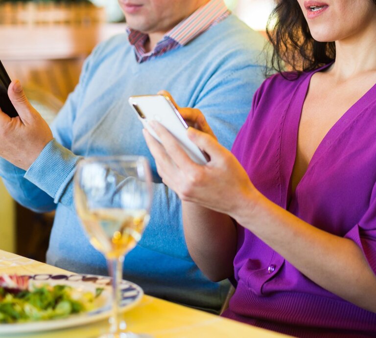 Eine Frau in lila Kleid und ein Mann in blauem Pulli starren beim Essen nebeneinander auf ihre Smartphones, ohne sich anzuschauen. | © Adobe Stock / JackF
