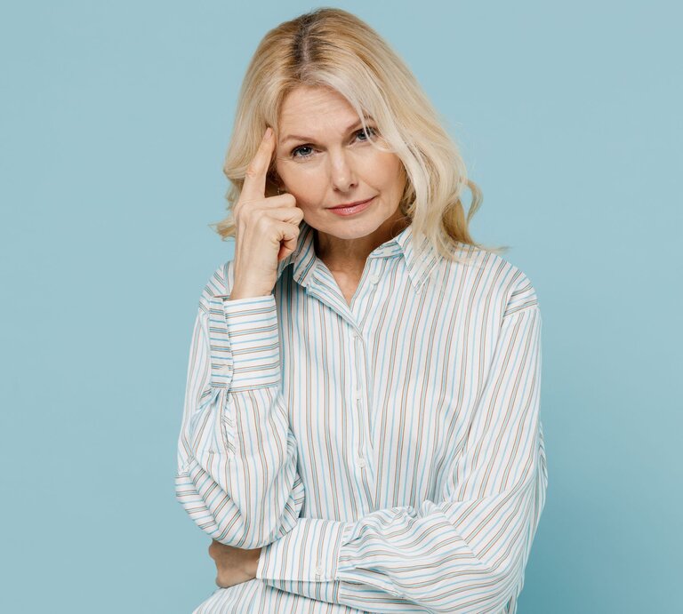 Blonde ältere Frau mit ernstem Blick tippt sich nachdenklich an die Schläfe vor hellblauem Hintergrund. | © Adobe Stock / ViDi Studio