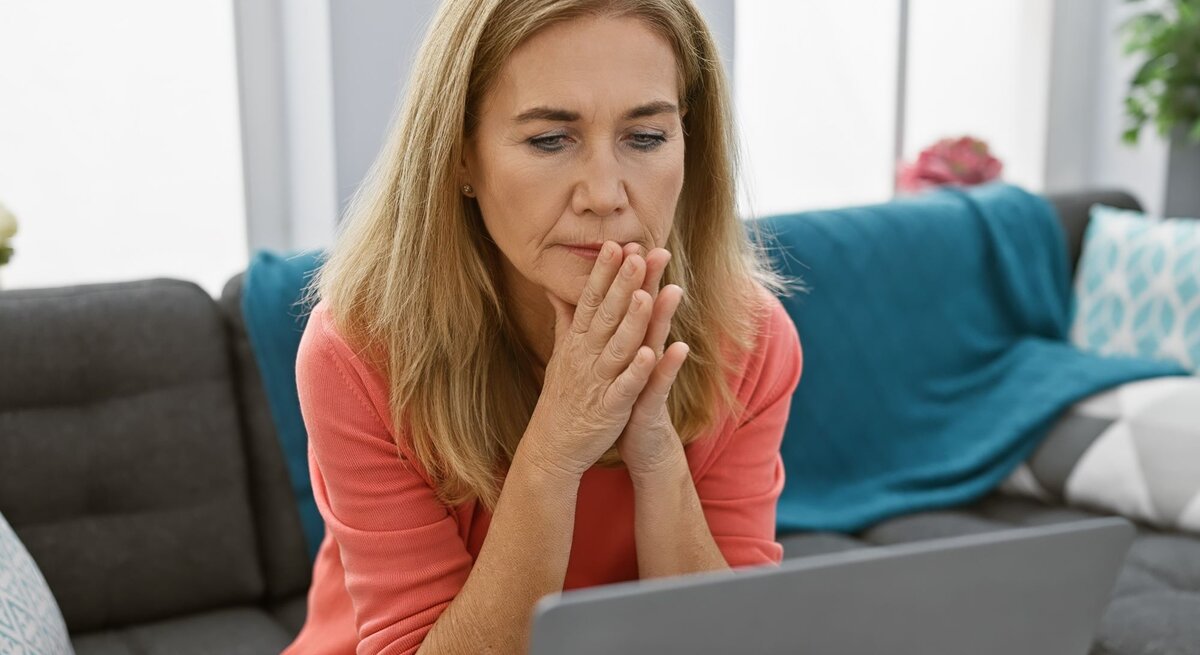 Eine nachdenkliche ältere Frau mit langen blonden Haaren sitzt im Wohnzimmer und blickt konzentriert auf ihren Laptop.