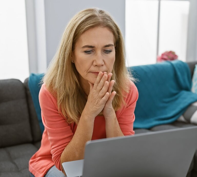 Eine nachdenkliche ältere Frau mit langen blonden Haaren sitzt im Wohnzimmer und blickt konzentriert auf ihren Laptop. | © Adobe Stock / Krakenimages.com