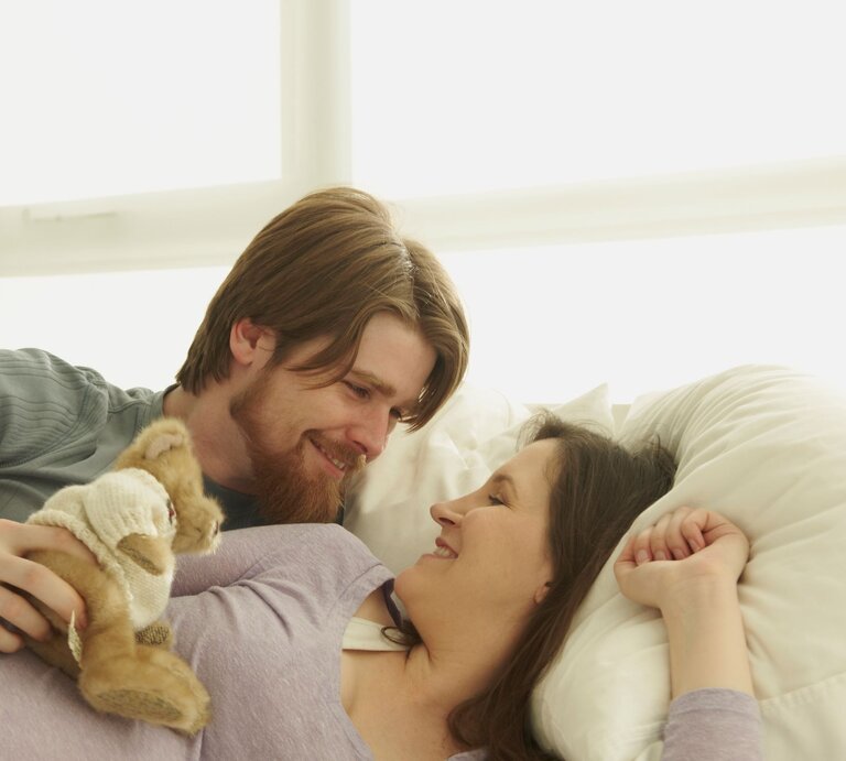 Ein lächelnder Mann mit braunem Bart und eine dunkelhaarige Frau in lila Oberteil liegen glücklich im Bett, mit einem Stofftier in seiner Hand. | © Getty Images