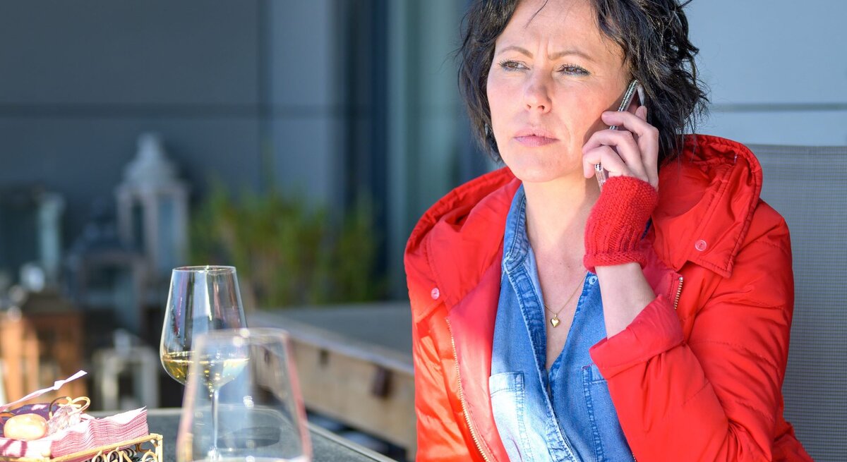 Frau, die in einem Restaurant sitzt und telefoniert. Sie trägt eine rote Jacke und wirkt nachdenklich. Auf dem Tisch vor ihr stehen zwei Weingläser, möglicherweise wartet sie auf jemanden.