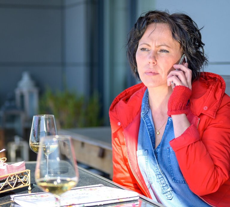 Frau, die in einem Restaurant sitzt und telefoniert. Sie trägt eine rote Jacke und wirkt nachdenklich. Auf dem Tisch vor ihr stehen zwei Weingläser, möglicherweise wartet sie auf jemanden. | © Adobe Stock / michaelheim
