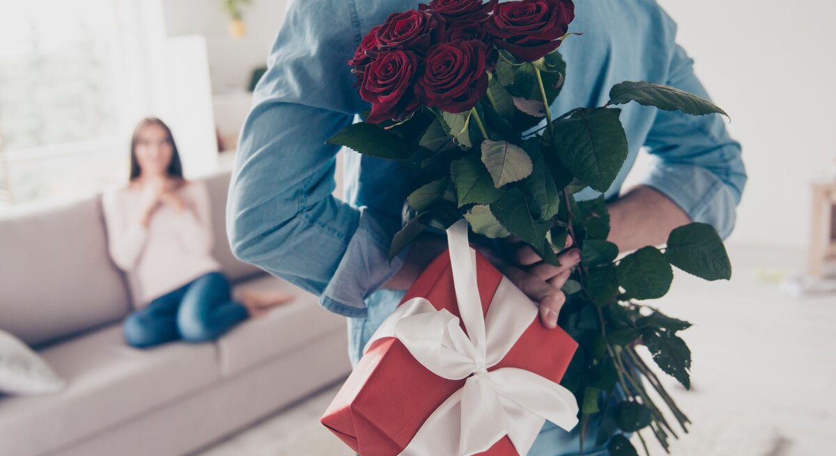 Junger Mann von hinten, der hinter seinem Rücken Rosen und ein Geschenk hält. Vor ihm ist schemenhaft eine Frau auf einem Sofa zu erkennen.