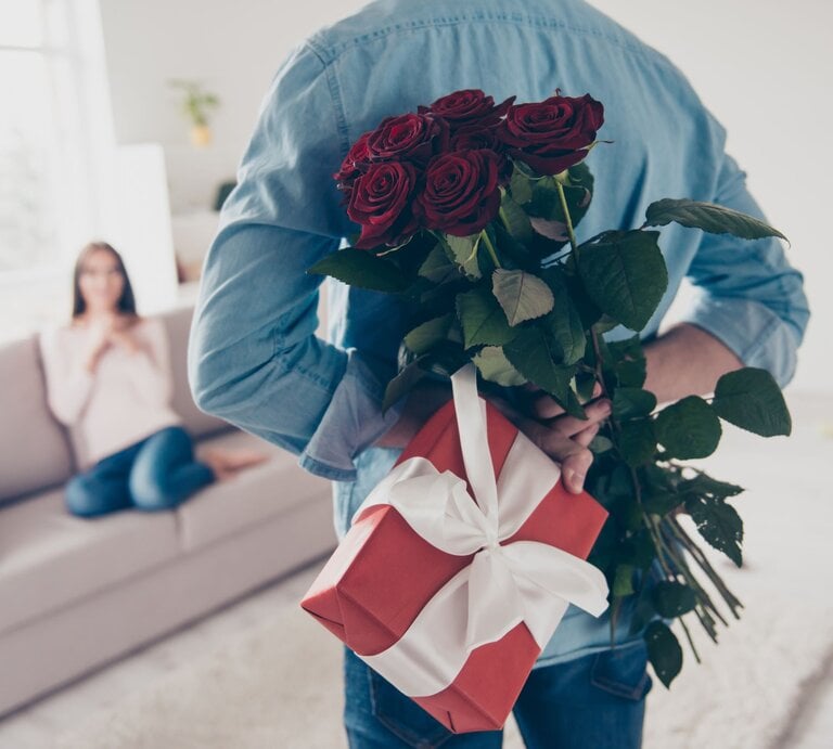 Junger Mann von hinten, der hinter seinem Rücken Rosen und ein Geschenk hält. Vor ihm ist schemenhaft eine Frau auf einem Sofa zu erkennen. | © Getty Images /Deagreez