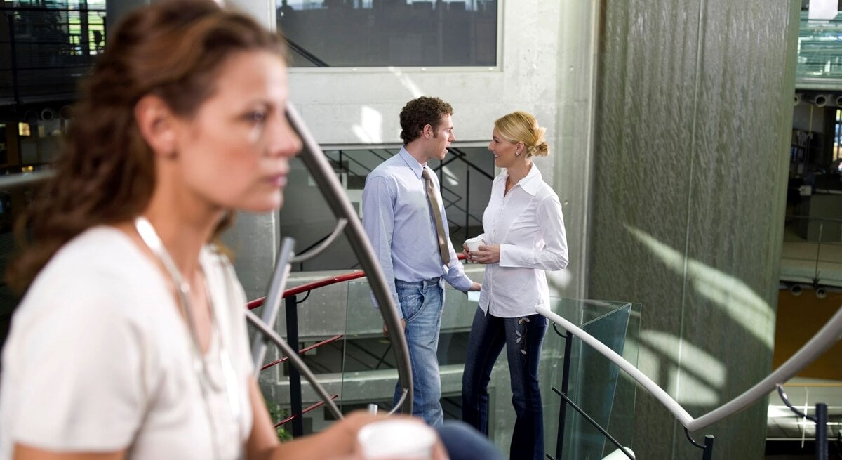 Eine Frau hört mit einem Getränk in der Hand am Arbeitsplatz einem Frau und einem Mann zu, die vertraulich wirken