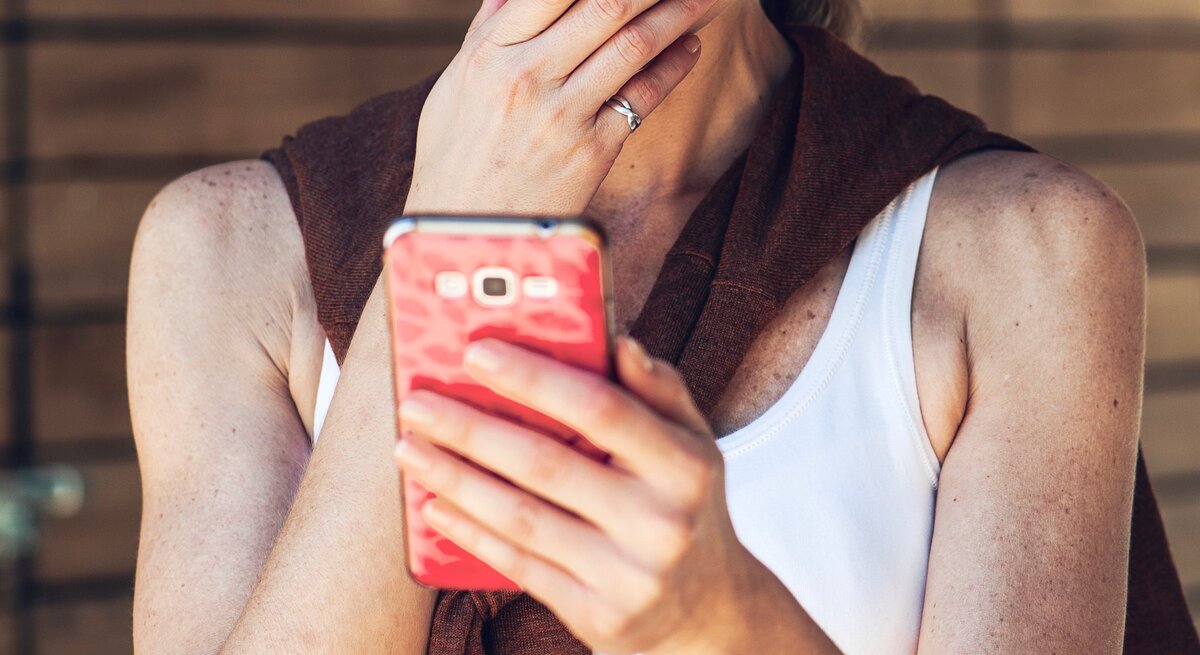 Eine Frau mit heller Haut und blondem Haar in weißem Top und braunem Pullover blickt nachdenklich auf ein pinkes Smartphone vor Holzwand.
