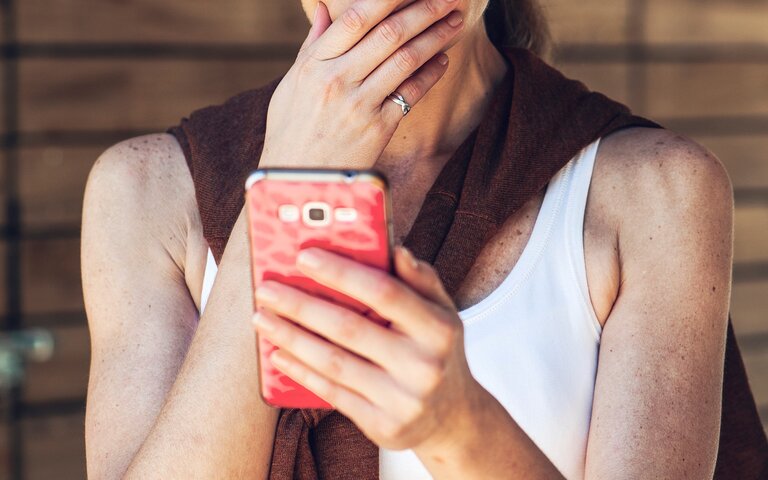 Eine Frau mit heller Haut und blondem Haar in weißem Top und braunem Pullover blickt nachdenklich auf ein pinkes Smartphone vor Holzwand. | © Getty Images / PhotoAlto/Frederic Cirou