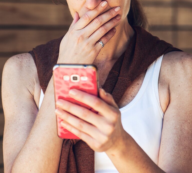 Eine Frau mit heller Haut und blondem Haar in weißem Top und braunem Pullover blickt nachdenklich auf ein pinkes Smartphone vor Holzwand. | © Getty Images / PhotoAlto/Frederic Cirou
