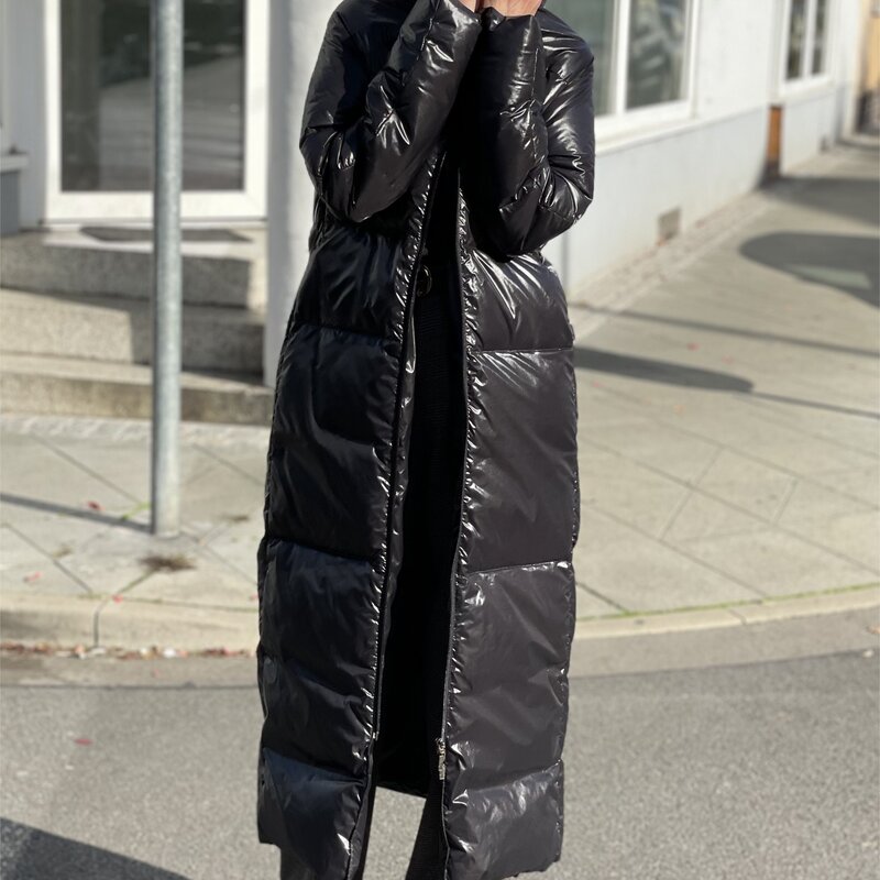 Frau in langem schwarzen Daunenmantel, grauer Hose und schwarzen Schuhen steht auf gepflastertem Weg, parkende Autos im Hintergrund. | © Renate Zott