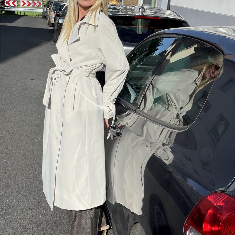 Frau in langem weißen Trenchcoat und beigefarbenen Schuhen lehnt seitlich an einem schwarzen Auto, sonnige Straße im Hintergrund. | © Renate Zott