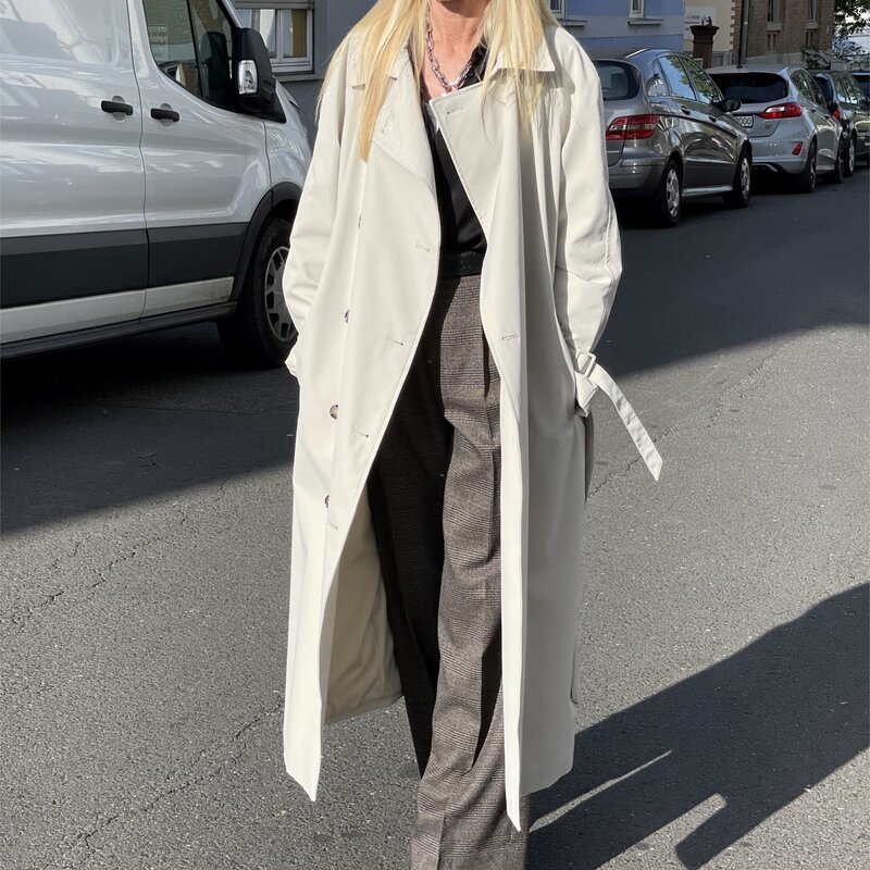 Frau mit langen blonden Haaren in weißem Trenchcoat, schwarzem Oberteil, grauer Hose und beigen Absatzschuhen auf sonniger Straße. | © Renate Zott