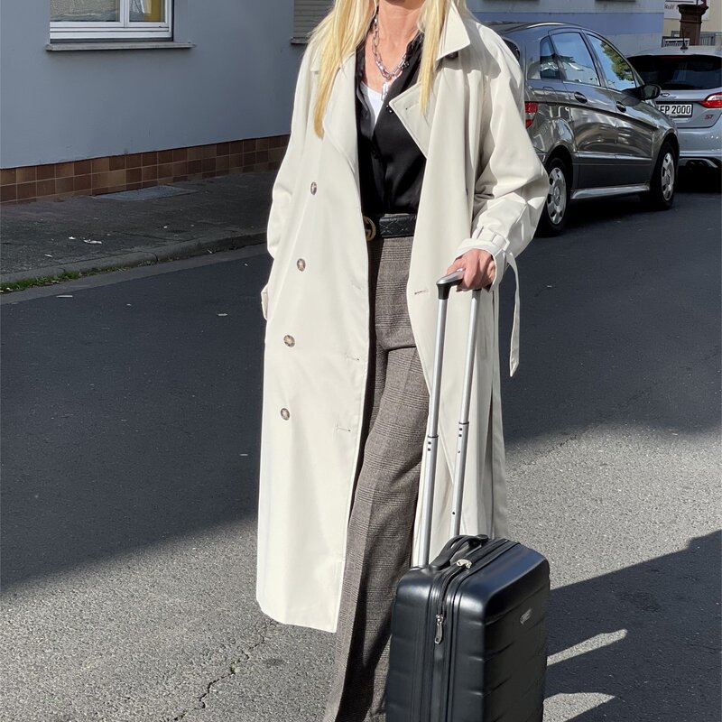 Frau in weißem Trenchcoat, schwarzem Oberteil und grauer Hose zieht einen schwarzen Rollkoffer auf einer Wohnstraße. | © Renate Zott