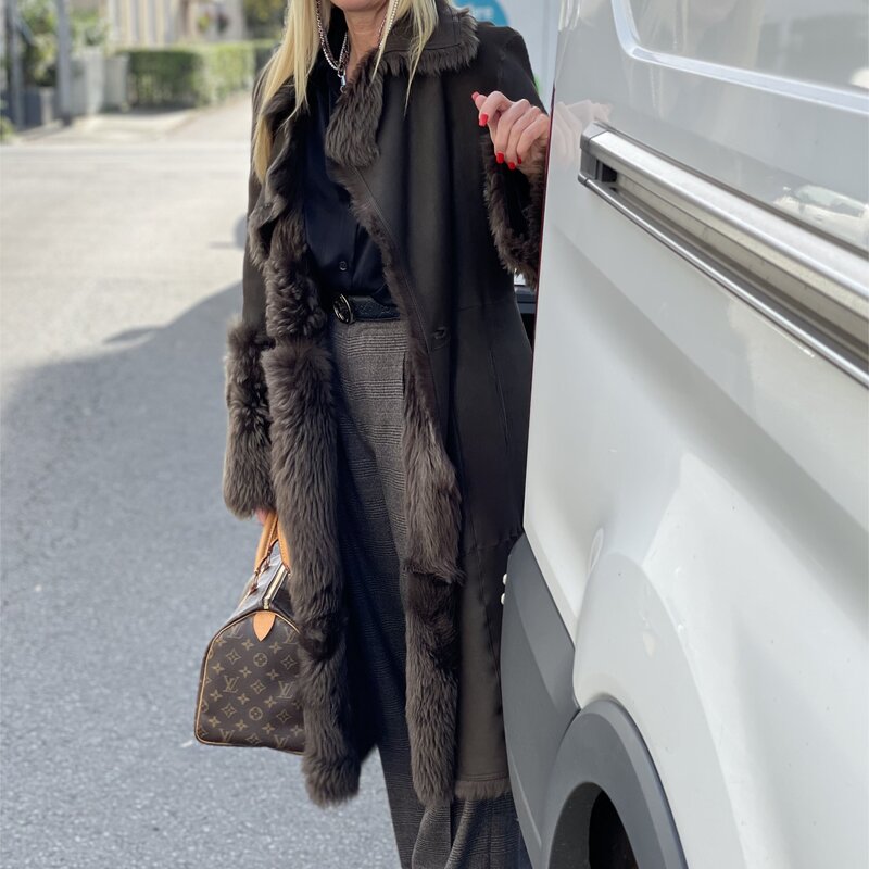 Frau in schwarzer Bluse, braunem Mantel mit Fellbesatz und grauer Hose lehnt an einem weißen Fahrzeug und hält eine Designer-Handtasche. | © Renate Zott