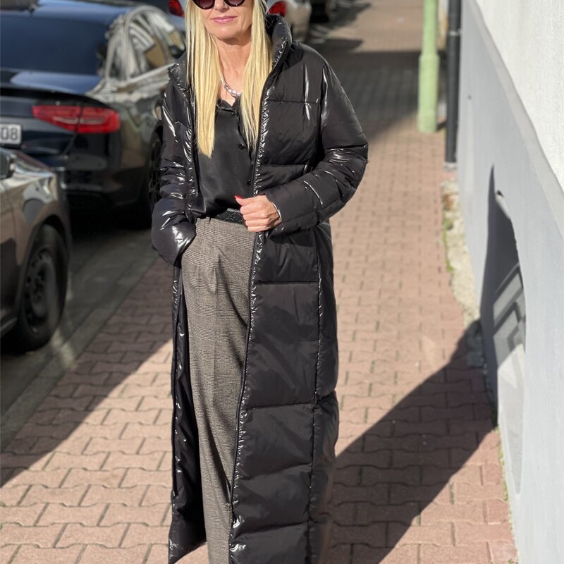 Frau in langem schwarzen Daunenmantel, grauer Hose und schwarzen Schuhen steht auf gepflastertem Weg, parkende Autos im Hintergrund. | © Renate Zott
