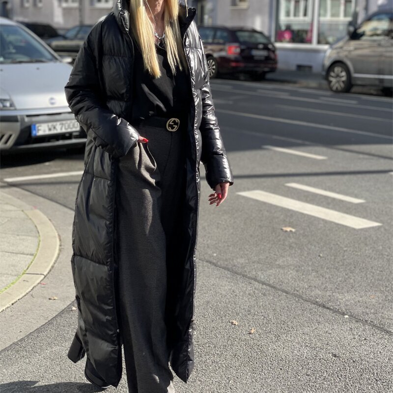 Frau in langem schwarzen Daunenmantel, grauer Hose und schwarzen Schuhen steht auf gepflastertem Weg, parkende Autos im Hintergrund. | © Renate Zott