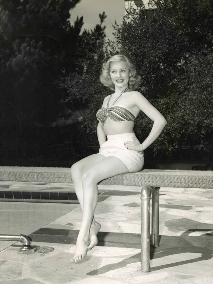 Blondhaarige Frau in Badeoutfit der 1950er-Jahre sitzt lächelnd am Rand eines Pools, umgeben von Bäumen. | © imago/Cola Images