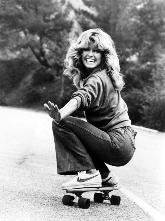 Junge Frau mit welligem Haar in Pullover und Schlaghose fährt lachend auf einem Skateboard auf einer Straße im Grünen. | © imago/ZUMA Press