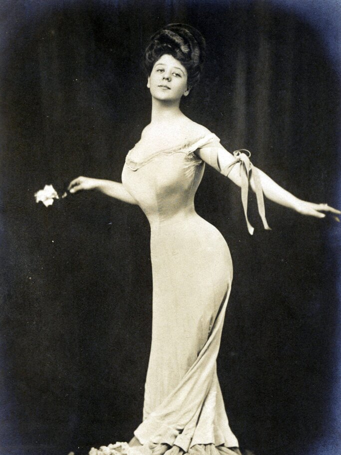 Junge Frau in figurbetontem Kleid mit betonter Taille, hält eine Blume und posiert graziös vor dunklem Hintergrund. | © imago/United Archives International
