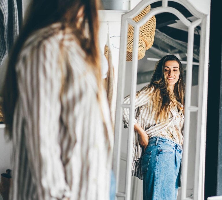 Junge Frau, die sich in einem großen, stehenden Spiegel betrachtet. Sie trägt eine lässige Jeans und eine gestreifte Bluse.  | © Adobe Stock / Mariia Korneeva