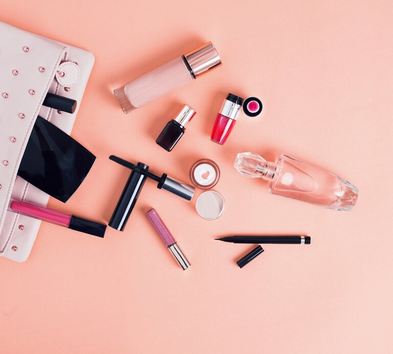 Ein flach angeordnetes Arrangement von Kosmetikprodukten auf einem pastellrosa Hintergrund. Zu den sichtbaren Gegenständen gehören eine hellrosa Kosmetiktasche mit Nieten, mehrere Lippenstifte und Lipglosse in verschiedenen Schattierungen, eine Wimperntusche, ein Flakon mit klarem Parfüm, ein Puderbehälter, ein flüssiges Make-up in einer Glasflasche, ein Nagellack und ein Augenbrauenstift. Die Anordnung ist sorgfältig platziert, um eine ästhetische Komposition zu schaffen, die sowohl Stil als auch die Vielfalt von Beauty-Produkten zeigt. | © iStock/AniDimi