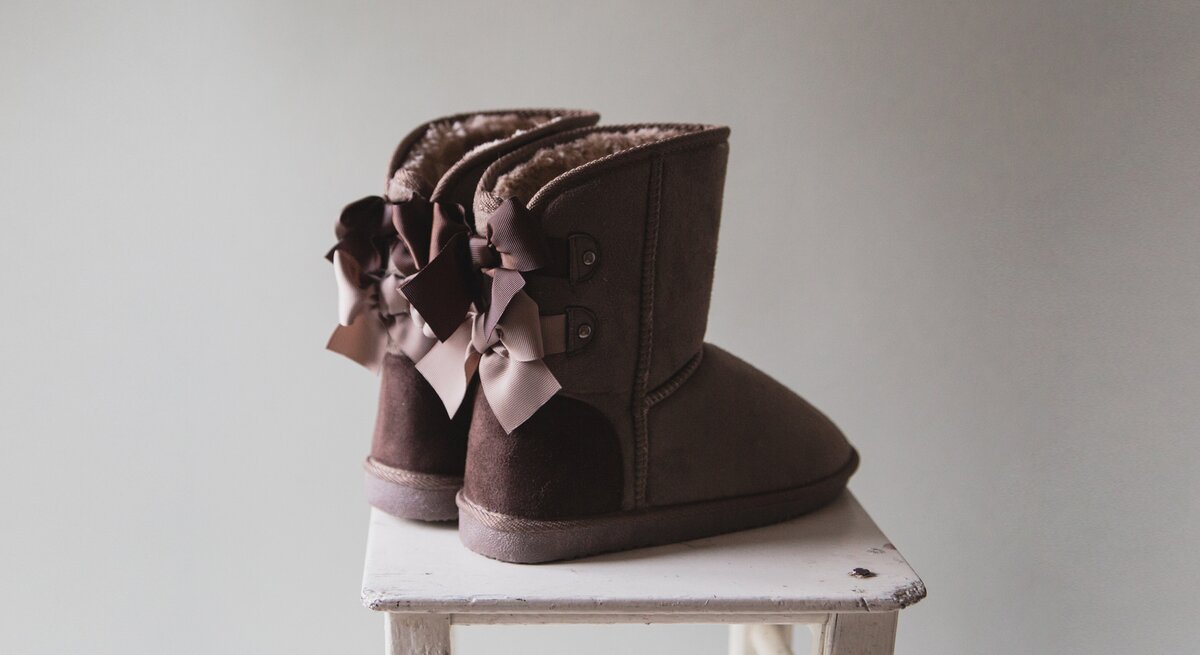 Ein Paar braune Ugg-Boots mit dekorativen Schleifen auf der Rückseite, die auf einem weißen, abgenutzten Holzhocker vor einem einfarbigen Hintergrund stehen.