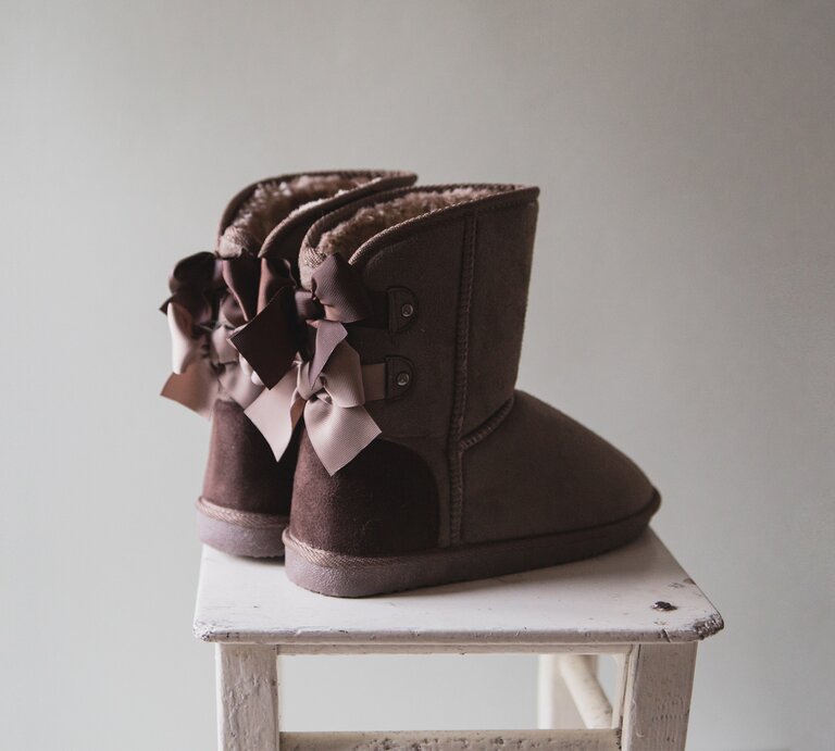 Ein Paar braune Ugg-Boots mit dekorativen Schleifen auf der Rückseite, die auf einem weißen, abgenutzten Holzhocker vor einem einfarbigen Hintergrund stehen. | © iStock/Bajneva