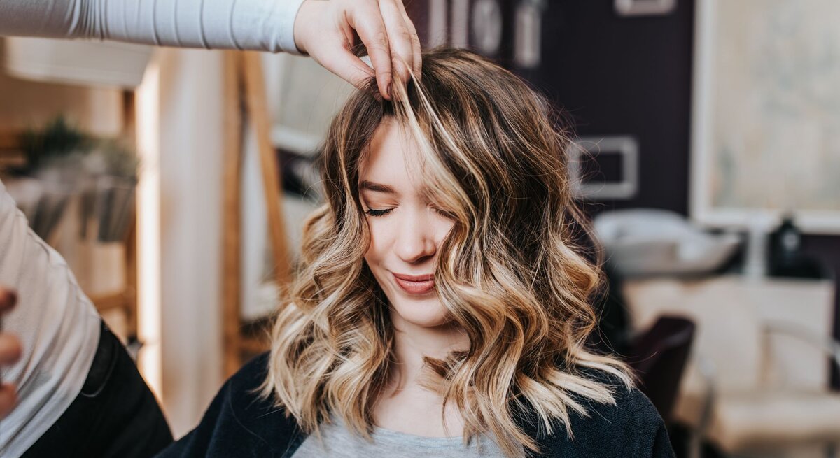 Junge Frau mit mittellangen, gewellten Haaren, die in einem Frisörsalon sitzt. Ihre Haare sind sorgfältig gestylt und weisen blonde Strähnen auf, die einen schönen Kontrast zu den dunkleren Haarpartien bilden. Eine Person, die nur teilweise und unscharf im Hintergrund zu erkennen ist, hebt mit den Fingern eine Strähne ihrer Haare an, vermutlich um die Frisur zu perfektionieren 
