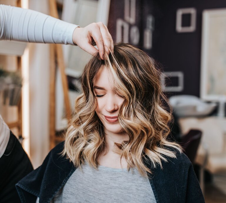 Junge Frau mit mittellangen, gewellten Haaren, die in einem Frisörsalon sitzt. Ihre Haare sind sorgfältig gestylt und weisen blonde Strähnen auf, die einen schönen Kontrast zu den dunkleren Haarpartien bilden. Eine Person, die nur teilweise und unscharf im Hintergrund zu erkennen ist, hebt mit den Fingern eine Strähne ihrer Haare an, vermutlich um die Frisur zu perfektionieren  | © Adobe Stock / hedgehog94