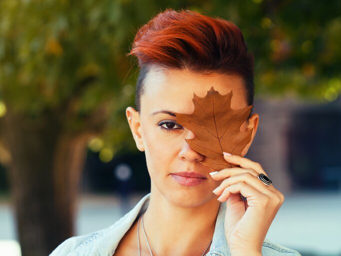 Frau mit roten Haaren trägt einen kurzen Undercut und hält ein Herbstblatt vor ihr Gesicht.  | © iStock/Elisaveta Ivanova