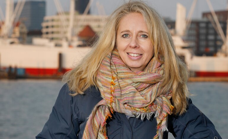 Eine lächelnde blonde Frau, bekleidet mit einem dunklen Wintermantel und einem farbenfrohen Schal, steht vor einer städtischen Flusskulisse mit modernen Gebäuden und Kränen im Hintergrund, was auf eine lebendige Hafenatmosphäre hindeutet. | © Sybill Schneider