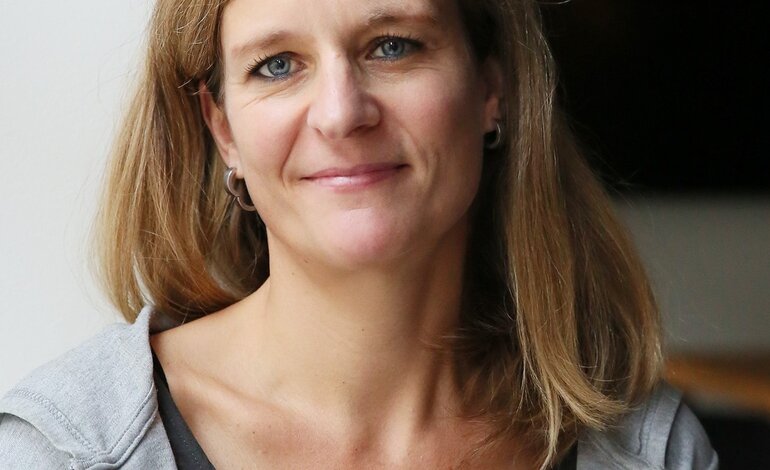 Das Bild zeigt eine Frau mit freundlichem Lächeln, die direkt in die Kamera blickt. Sie hat mittellanges blondes Haar, blaue Augen und trägt ein graues T-Shirt mit einer offenen grauen Jacke. Der Hintergrund ist schlicht und unauffällig, was den Fokus auf die Frau lenkt. Ihr Ausdruck wirkt offen und zugänglich. | © BILD der FRAU