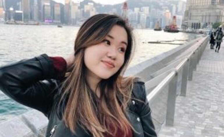 Eine junge Frau mit langem, welligem Haar und einer schwarzen Lederjacke steht lächelnd am Ufer einer Hafenpromenade. Sie hat eine Hand leicht in ihrem Haar und blickt träumerisch zur Seite. Im Hintergrund ist eine Stadtlandschaft mit Hochhäusern und der Silhouette eines bewölkten Himmels über einer Wasserfläche zu erkennen. | © privat