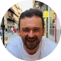 Das Bild zeigt einen lächelnden Mann mit kurzen dunklen Haaren und Bart. Er trägt ein weißes T-Shirt und sitzt im Freien, wie es scheint, in einem Straßencafé. Im Hintergrund ist eine belebte Straße mit historischen Gebäuden zu erkennen. | © privat