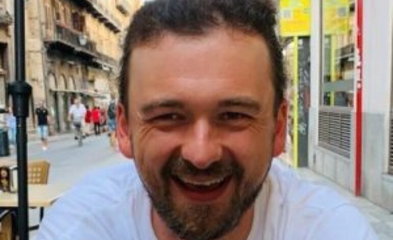 Das Bild zeigt einen lächelnden Mann mit kurzen dunklen Haaren und Bart. Er trägt ein weißes T-Shirt und sitzt im Freien, wie es scheint, in einem Straßencafé. Im Hintergrund ist eine belebte Straße mit historischen Gebäuden zu erkennen. | © privat