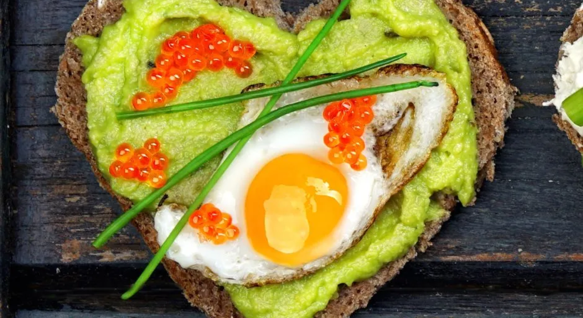 Herzförmiges Vollkornbrot mit Avocado-Creme, Spiegelei, rotem Kaviar und Schnittlauch auf dunkler Holzunterlage – gesundes, eiweißreiches Frühstück.