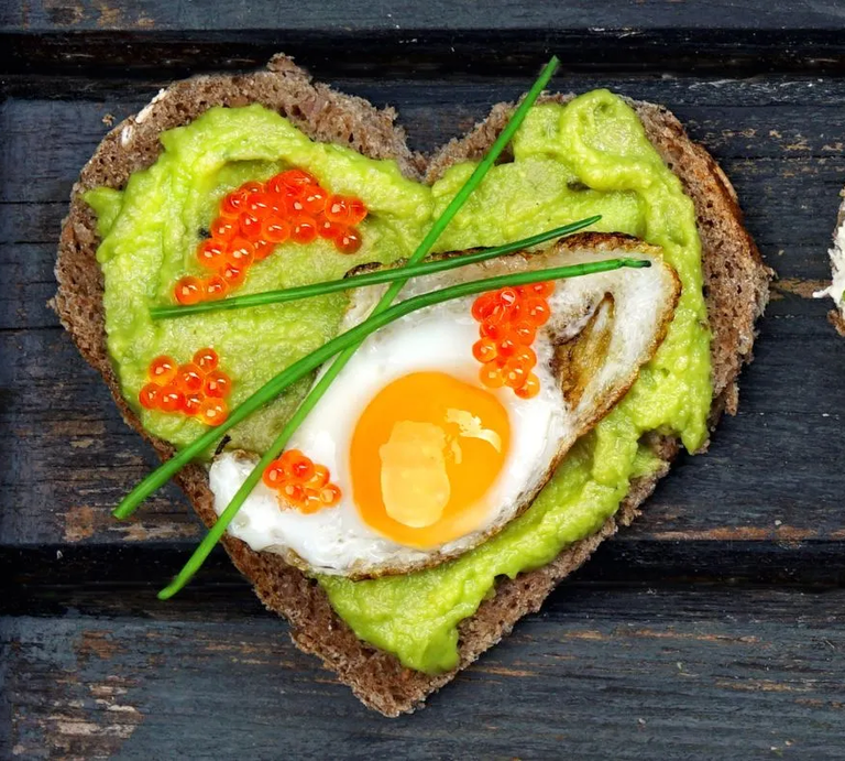 Herzförmiges Vollkornbrot mit Avocado-Creme, Spiegelei, rotem Kaviar und Schnittlauch auf dunkler Holzunterlage – gesundes, eiweißreiches Frühstück. | © Adobe Stock/ Jenny Sturm