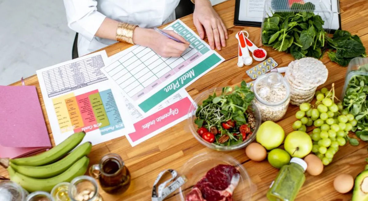 Ernährungsberaterin erstellt einen Ernährungsplan am Tisch, umgeben von frischen Lebensmitteln wie Obst, Gemüse, Fleisch, Nüssen und Vollkornprodukten sowie Unterlagen zu Makronährstoffen und einem Maßband.