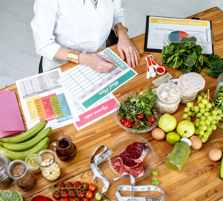Ernährungsberaterin erstellt einen Ernährungsplan am Tisch, umgeben von frischen Lebensmitteln wie Obst, Gemüse, Fleisch, Nüssen und Vollkornprodukten sowie Unterlagen zu Makronährstoffen und einem Maßband. | © RossHelen via Canva.com