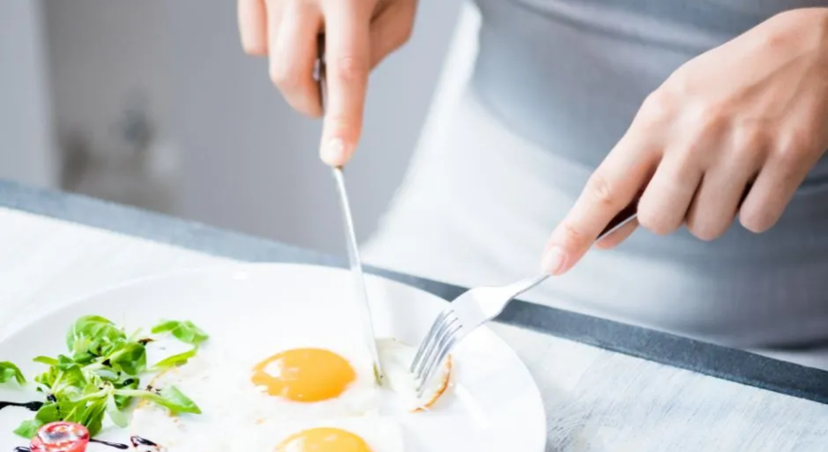 Person schneidet zwei Spiegeleier auf einem Teller an, serviert mit etwas grünem Salat, einer halben Kirschtomate und Balsamico-Dekor, auf einem hellen Tisch – Nahaufnahme einer Mahlzeit.