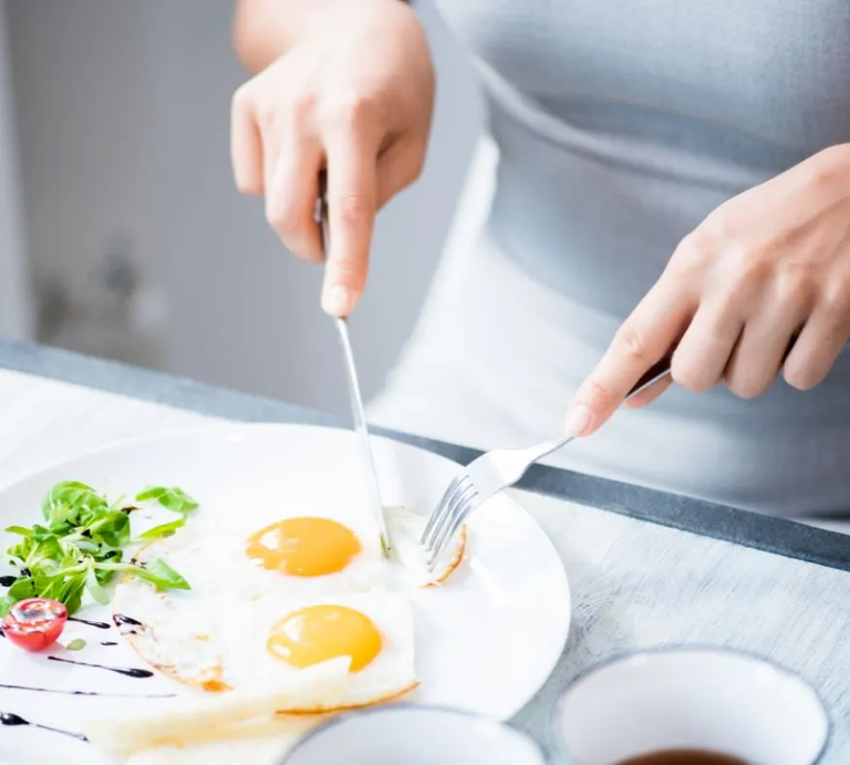 Person schneidet zwei Spiegeleier auf einem Teller an, serviert mit etwas grünem Salat, einer halben Kirschtomate und Balsamico-Dekor, auf einem hellen Tisch – Nahaufnahme einer Mahlzeit. | © Getty Images/ SeventyFour