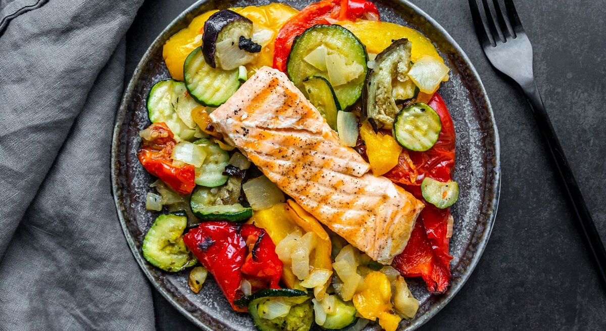 Das Bild zeigt einen Teller mit einer gesunden Mahlzeit, bestehend aus einem Stück gegrilltem Lachs auf einem Bett aus verschiedenen gegrillten oder gebratenen Gemüsen. Die Gemüsesorten scheinen Zucchini, Paprika und Zwiebeln zu umfassen