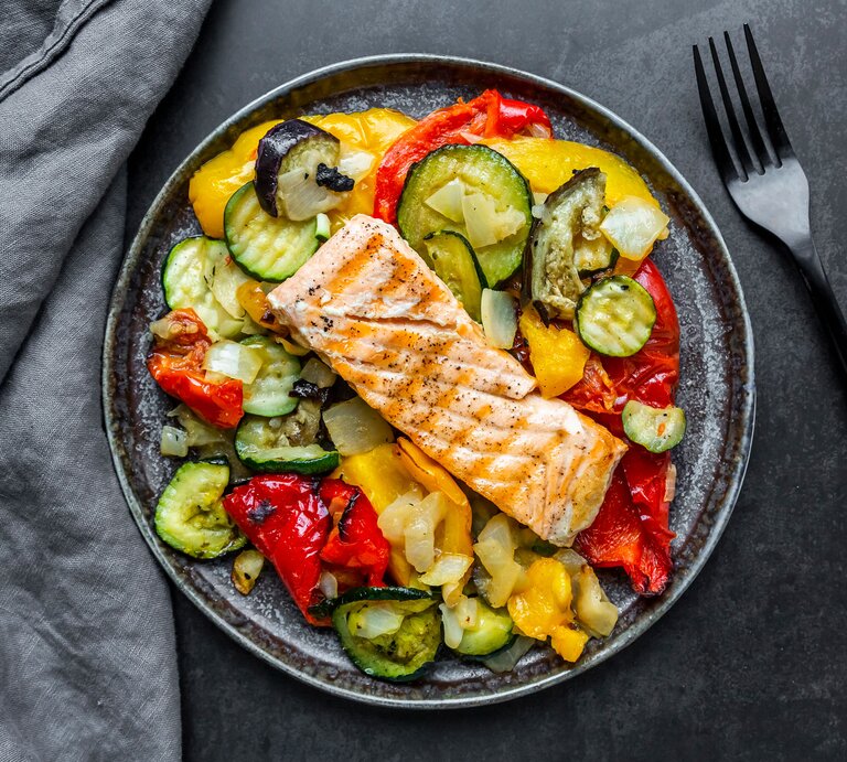 Das Bild zeigt einen Teller mit einer gesunden Mahlzeit, bestehend aus einem Stück gegrilltem Lachs auf einem Bett aus verschiedenen gegrillten oder gebratenen Gemüsen. Die Gemüsesorten scheinen Zucchini, Paprika und Zwiebeln zu umfassen | © Getty-Images/lacaosa